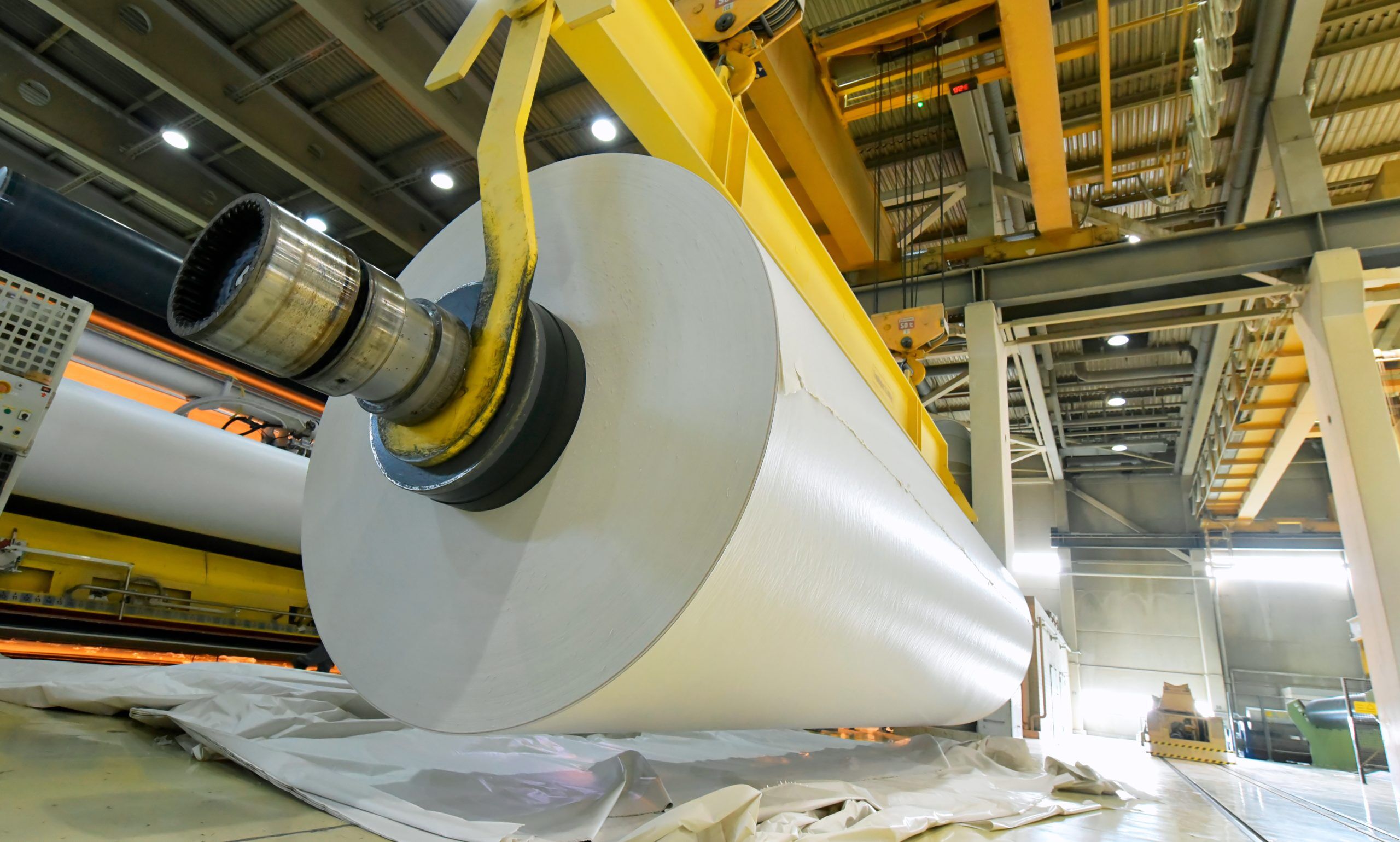 Industrial sized roll of paper on yellow crane next to paper press