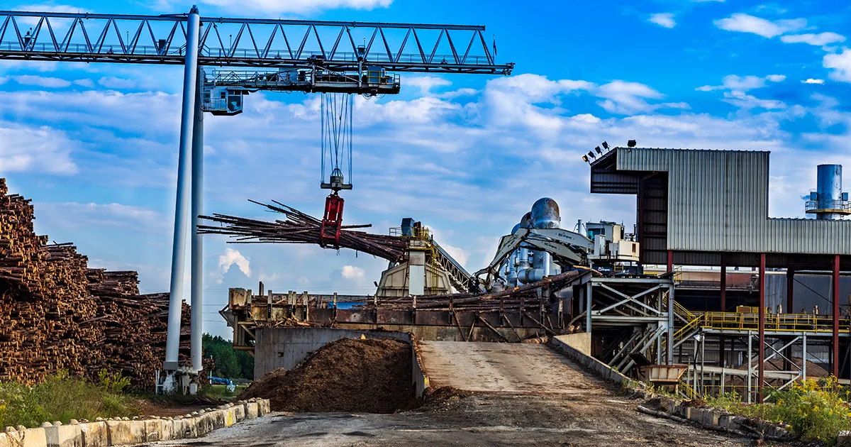 Crane at a wood processing facility lifting a bundle of wooden poles over a yard with stacked timber and industrial equipment nearby